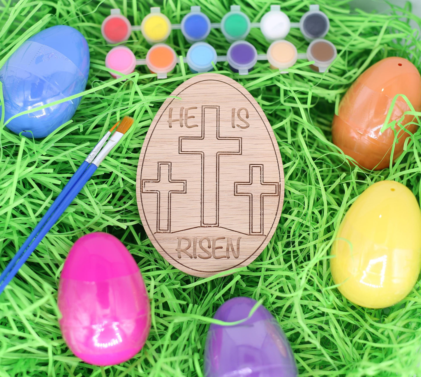 Wooden Easter egg with 'He is Risen' message surrounded by colorful plastic eggs on green grass.