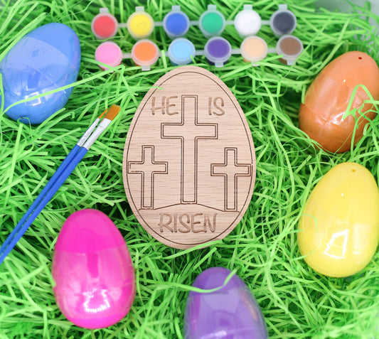 Wooden Easter egg with 'He is Risen' message surrounded by colorful plastic eggs on green grass.